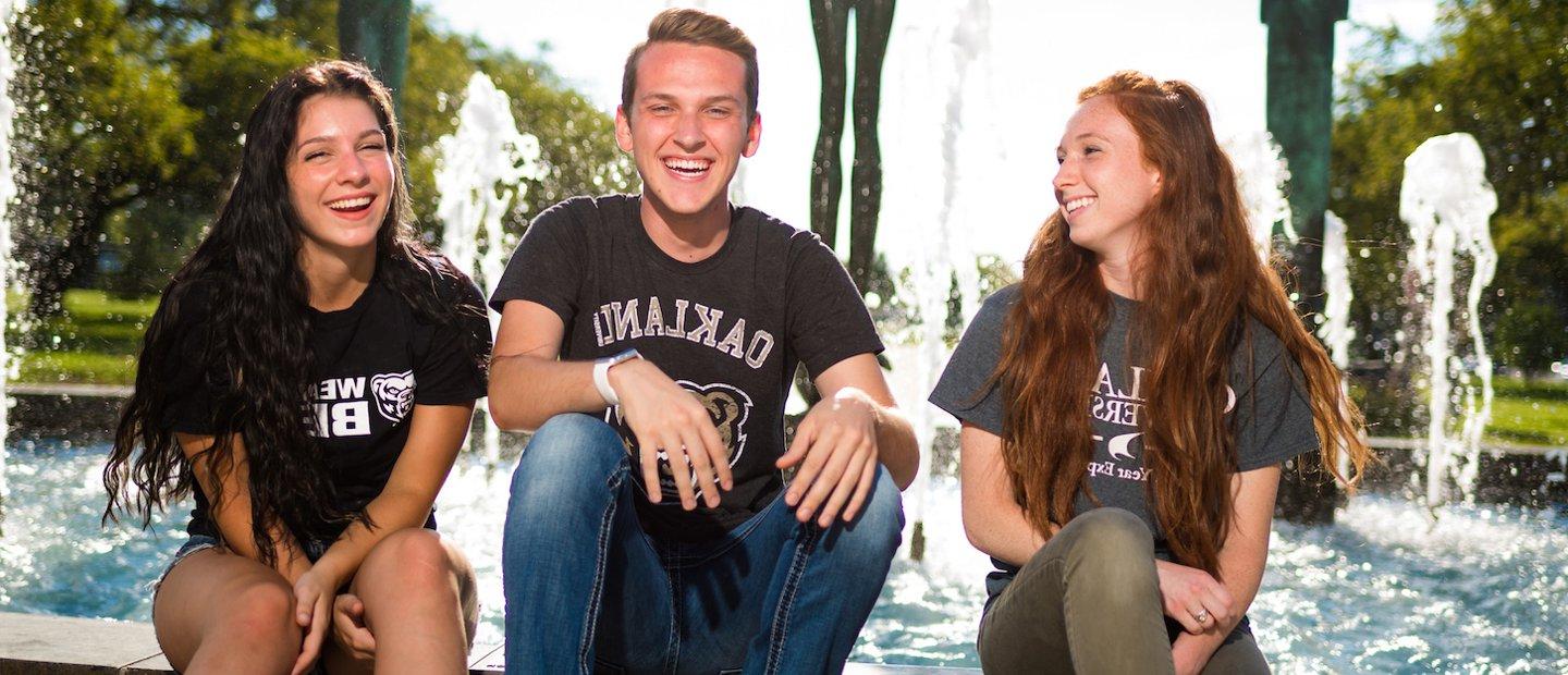 Three Oakland University students sitting on the edge of a fountain, laughing and smiling.