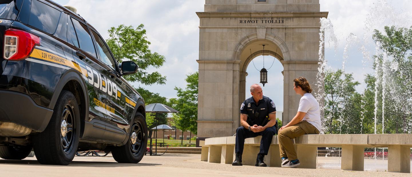 A police officer sitting with a person on a bench in front of Elliott Tower.