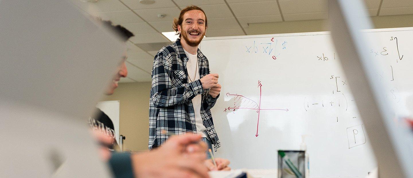 A tutor standing in front of a white board with mathematical equations, teaching a student.