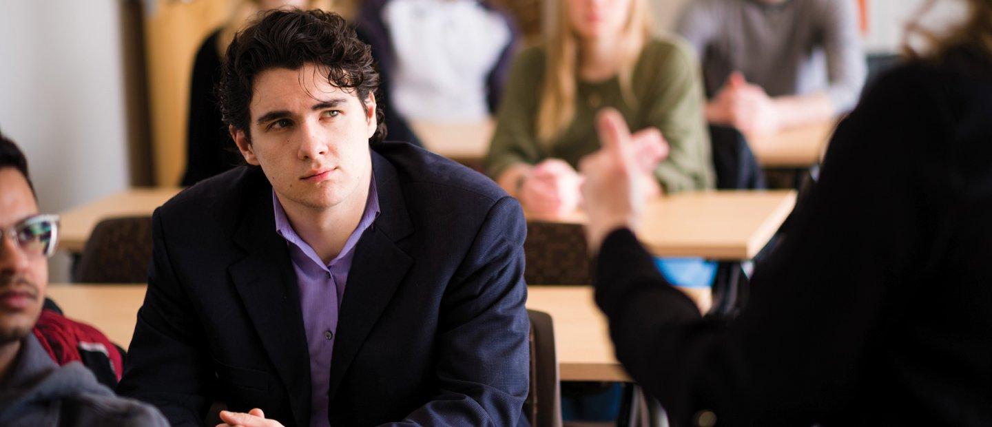 young man in a classroom full of students, watching someone at the front of the room