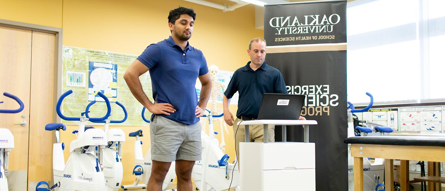 A man standing on a scale while another man reads data on a computer screen, in a fitness studio.