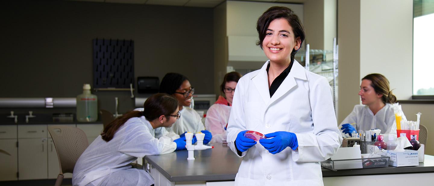 young woman in a white lab coat and blue gloves, smiling at the camera