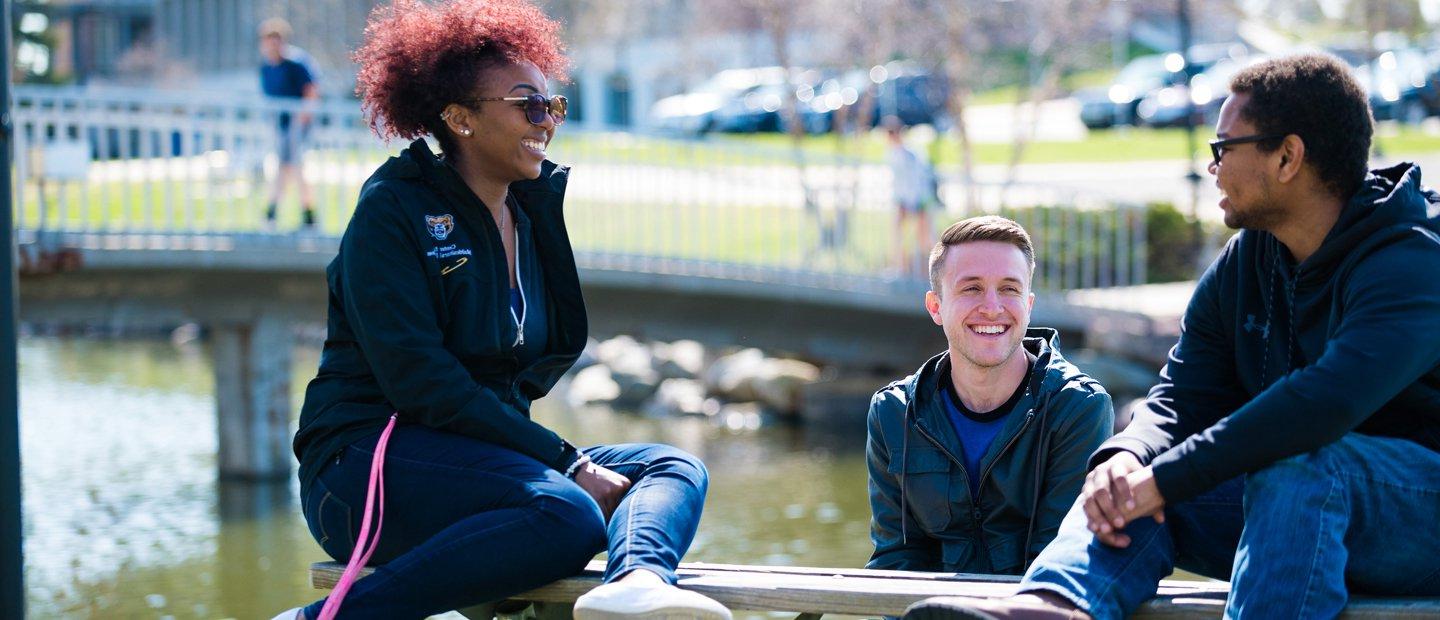 two male and one female O U student seated outside in front of a lake with a bridge in the background